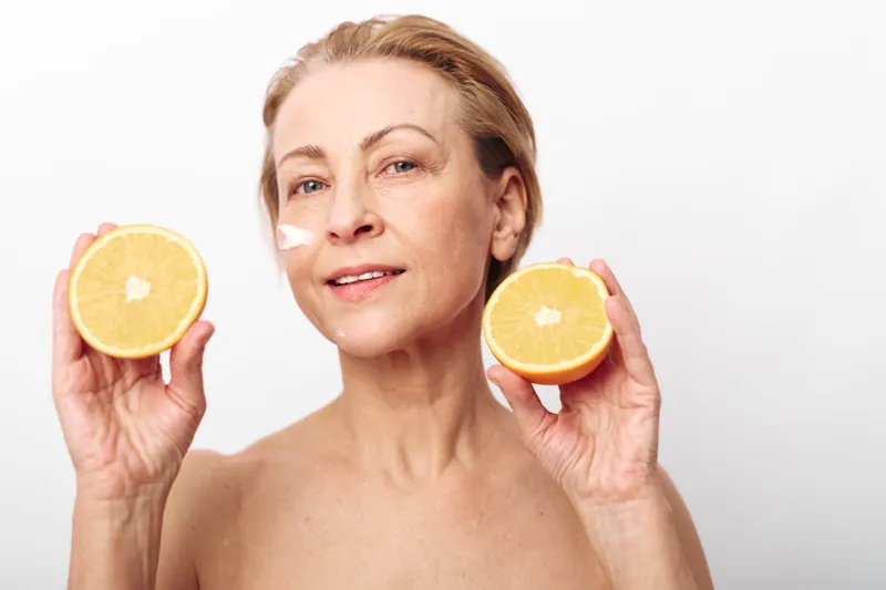 Mature woman with natural skin holds two halves of a fresh orange, showing facial cream on cheek. Concept for skincare, natural beauty, wellness, and healthy lifestyle with citrus fruit.