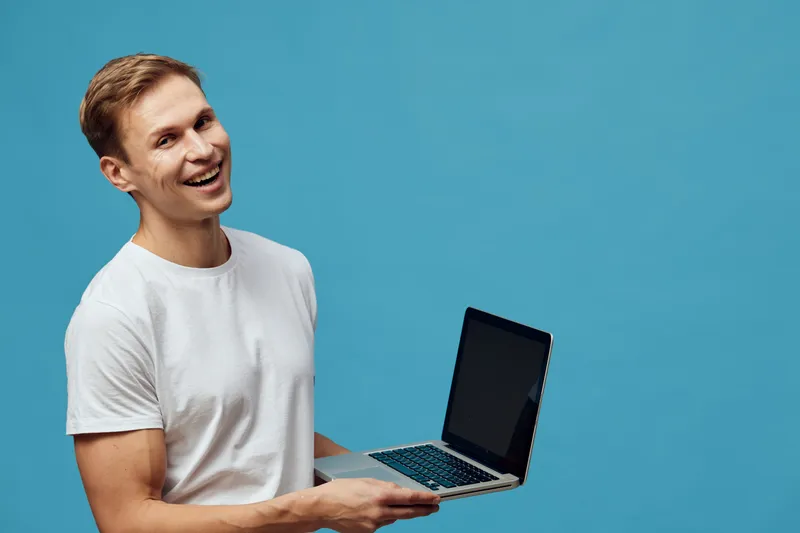 Smiling young man holding a laptop in hand, wearing casual white t-shirt, standing against a plain blue background. Happy, cheerful, modern, technology, lifestyle, studio portrait.