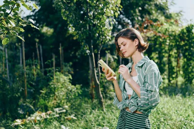 Young woman stands in a sunlit park, focused on her smartphone while holding a bottle, enjoying a peaceful outdoor moment.