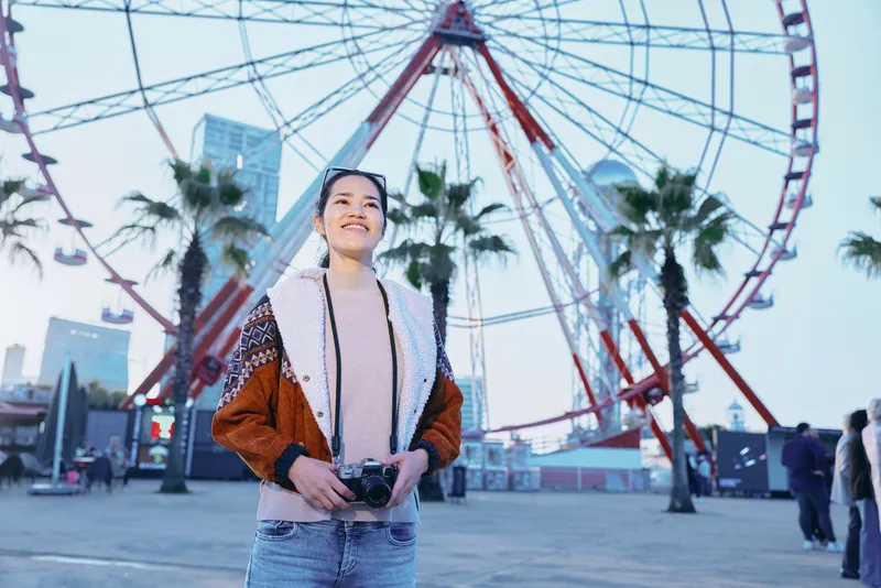 Woman with camera on a boardwalk near a towering ferris wheel, palm trees and city skyline, capturing a lively outdoor travel moment under airy daylight and soft urban breeze