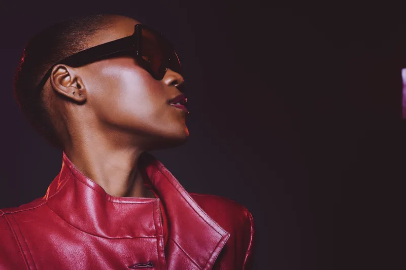 Stylish black woman with dark skin wearing oversized sunglasses and a red leather jacket looks sideways on black background. Concept of fashion, confidence, and modern urban style.