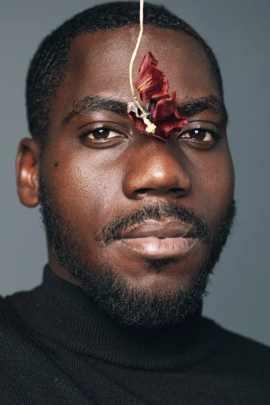 African man with dark skin and beard in creative conceptual style wearing black turtleneck with dried flower hanging on face for artistic portrait and visual expression.
