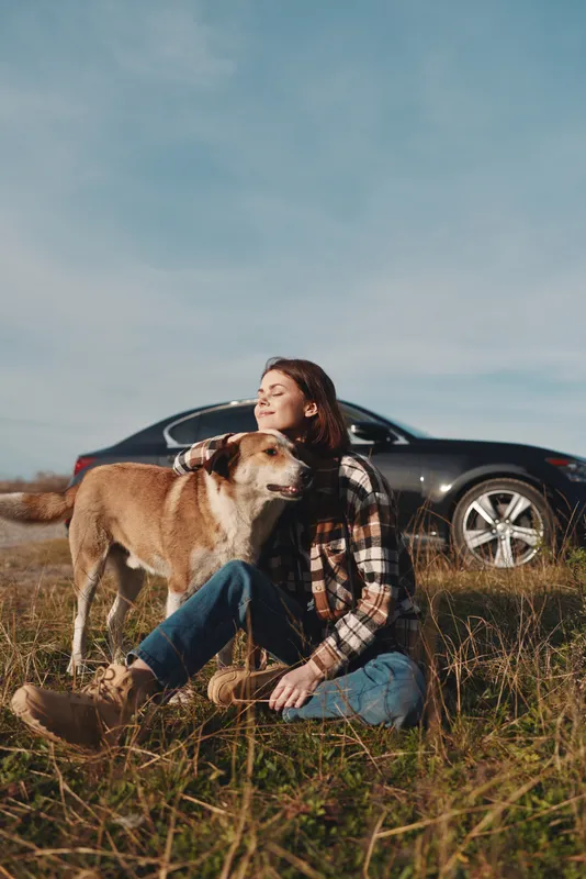 Woman sitting in a field with her dog, relaxing outdoors beside a car under a clear sky. Casual plaid shirt and jeans, a peaceful moment of companionship and nature on a roadside escape