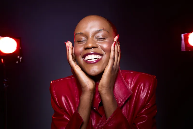 Happy dark skinned African woman in red leather jacket smiles with closed eyes and hands on face. Studio portrait with dramatic lighting and joyful mood. Concept of beauty and confidence.