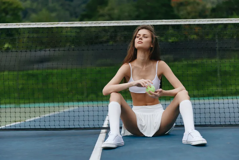 Young athletic woman enjoying healthy snack on tennis court, wearing sports attire and seated in front of net in summer setting