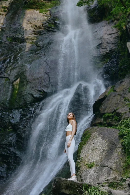 Young woman standing near a powerful waterfall in white activewear, surrounded by rocky cliffs and lush greenery, enjoying nature and outdoor adventure.