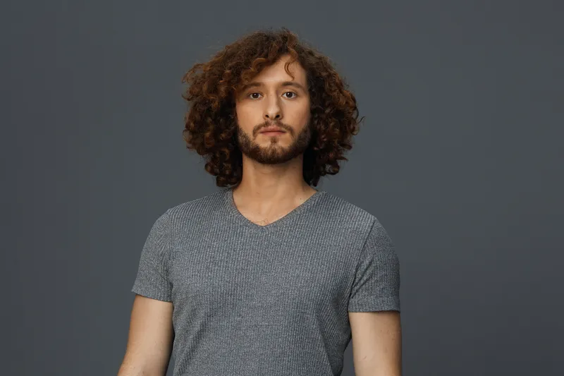 Casual portrait of a young man with curly hair in a gray T shirt against a neutral background, expressing confidence and tranquility, ideal for lifestyle and personal branding