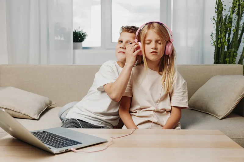 Two children share pink headphones while sitting on a beige couch in a bright living room with a laptop on the table. Concept of sibling bonding and technology.