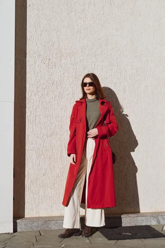 Stylish woman in a red trench coat stands against a sunlit textured wall, wearing sunglasses and beige wide?leg pants, exuding confident urban chic.