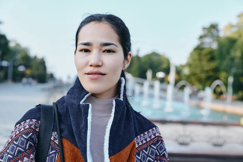 Woman outdoor jacket fashion street park fountain urban setting with patterned coat, thoughtful gaze, candid urban style, showcasing street fashion in daylight near a water feature