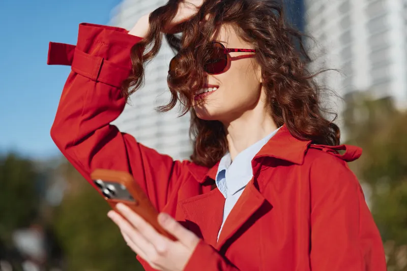 Young woman in a red coat checks her phone with a hopeful smile against a blurred urban background, the sunlight highlighting her silhouette