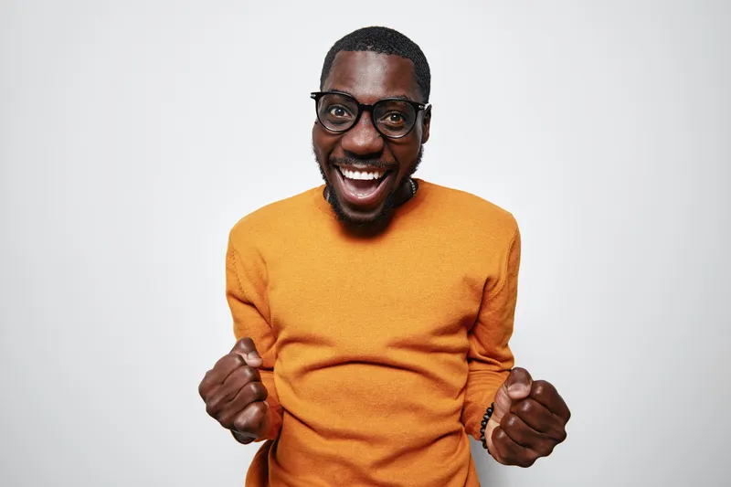African American man with dark skin wearing glasses and orange sweater smiling excitedly with clenched fists on white background. Happy, success, joy and celebration concept.