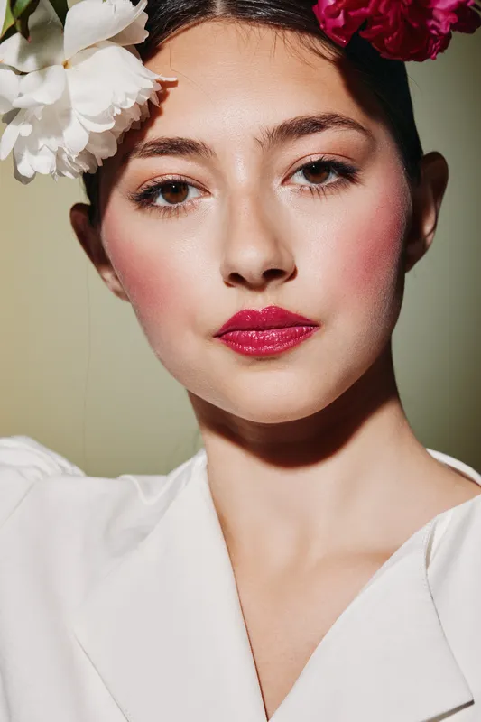 Closeup of beauty model with bright floral hairpiece, glossy fuchsia lips, and flawless radiant skin wearing white dress. Avant-garde makeup in creative fashion concept.
