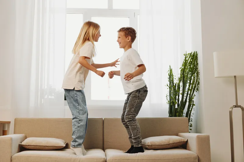 Two happy children jump on beige sofa in bright living room with large window and green plant. Concept of childhood joy, playful siblings, and indoor family fun activities.