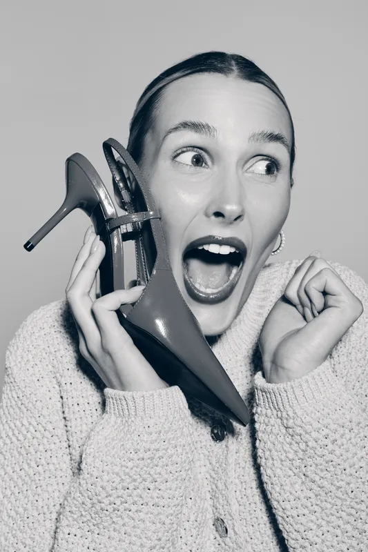 Caucasian woman with light skin holds high heel shoe near her face with surprised expression. Studio portrait in black and white. Concept for fashion, emotions, and footwear.