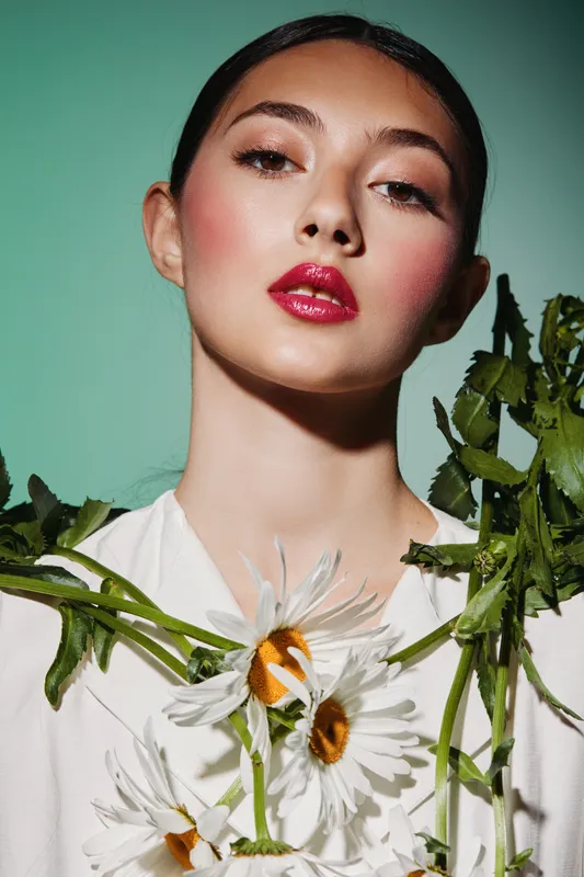Portrait of creative young woman with glossy red lips and natural flowers on white shirt against green background. Concepts of beauty fashion and floral style.