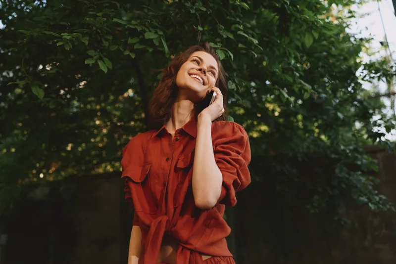 A woman in a red dress chats on her phone outdoors, smiling among trees, capturing a candid lifestyle portrait in natural light