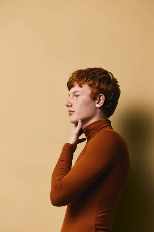 Young man with red hair and fair skin wearing brown turtleneck posing in profile against a beige background with thoughtful facial expression and hand on chin.