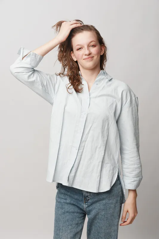 Young woman with wet hair wearing casual shirt and jeans smiles gently while touching her head. Studio portrait on white background. Concept of natural beauty and relaxed mood.