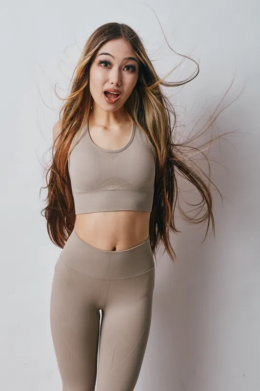 Young woman in beige sportswear with long flowing hair stands surprised against white wall. Female athlete or fitness model posing in casual activewear with natural expression indoors.
