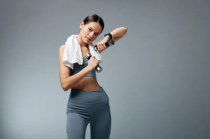 Woman in athletic wear with towel around neck, wiping sweat while lifting a dumbbell, mid workout in a studio. Focused fitness, strength, and momentum for sport training.