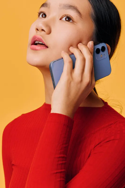 Woman talking on smartphone with thoughtful expression against yellow background. Female holding phone near ear wearing red sweater in casual communication and lifestyle scene.