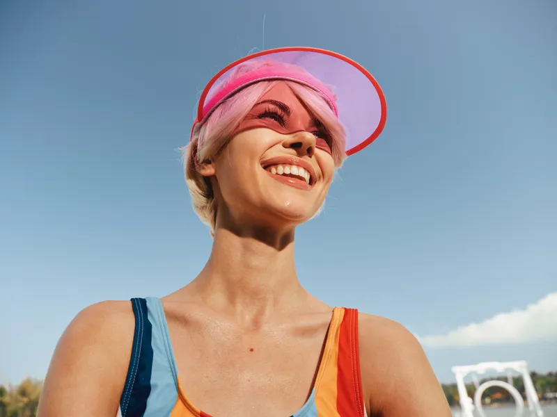 A joyful young woman wearing a pink visor and colorful workout outfit smiles under a clear blue sky, exuding vibrant energy and carefree summer fitness.