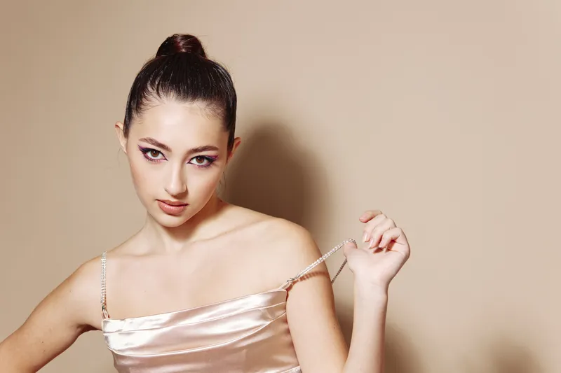 Young woman in shiny satin dress pulls delicate strap off her shoulder, posing creatively against neutral background. Concept of beauty, fashion, satin fabric, and style.