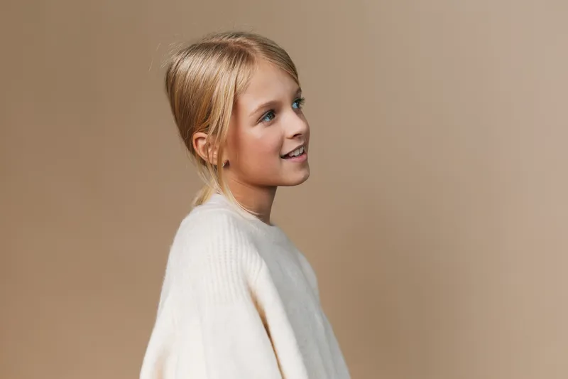 Portrait of a young girl in a soft knit sweater, gazing to the side with a gentle smile. Warm neutral backdrop and soft lighting highlight innocence, calm, and curiosity.