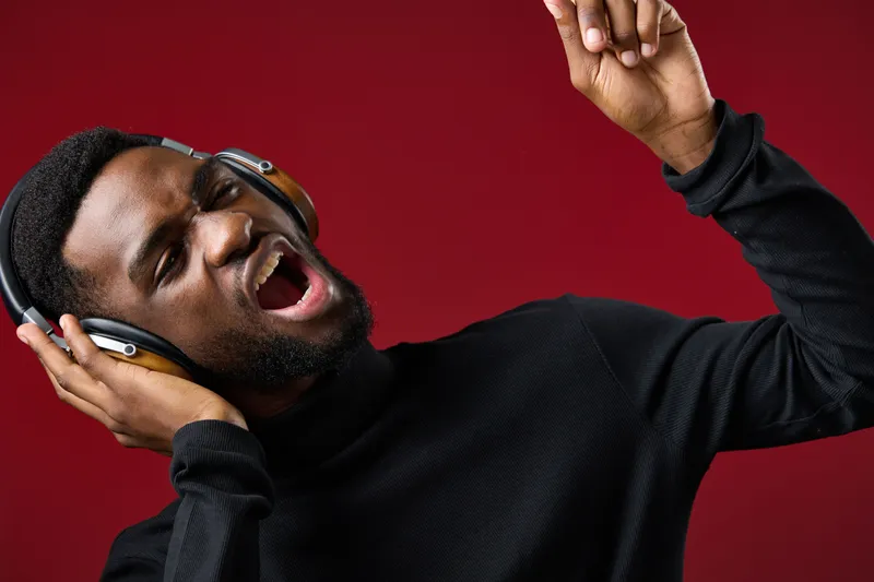 Young African man singing passionately with headphones on a red background, expressing joy and enthusiasm for music in a stylish black outfit