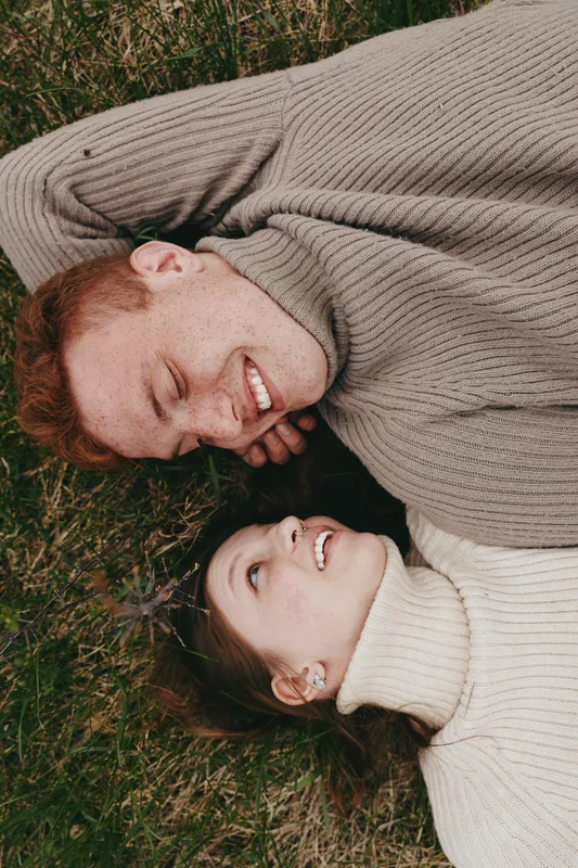 Young redhead man and brunette woman lie on grass looking at each other smiling in cozy turtleneck sweaters enjoying a relaxed moment outdoors in autumn lifestyle setting.