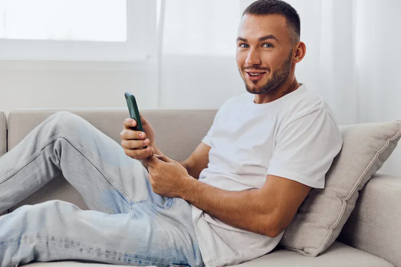 A smiling man engaged in a phone call, showcasing emotional vulnerability while sitting comfortably at home in a cozy setting. Perfect for modern therapy culture.