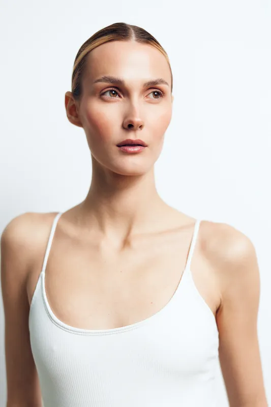 Young woman with blonde hair wearing a white tank top stands against a plain white backdrop, looking off to the side with a calm, thoughtful expression.