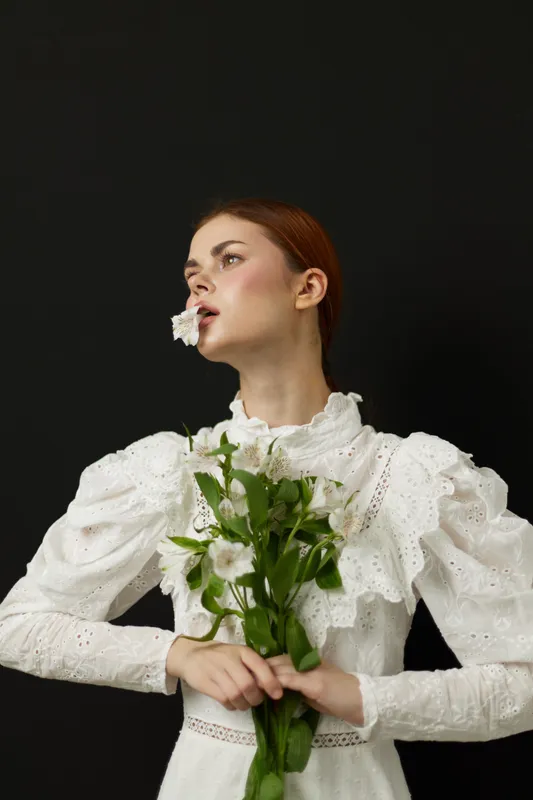 Elegant woman in a white dress holding flowers, showcasing beauty and serenity against a dark background, ideal for nature and fashion related projects