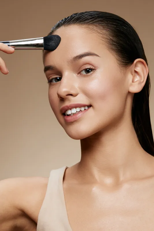 Young woman applying makeup with a brush on her forehead, showcasing a natural, radiant beauty against a soft beige background ideal for beauty and skincare concepts