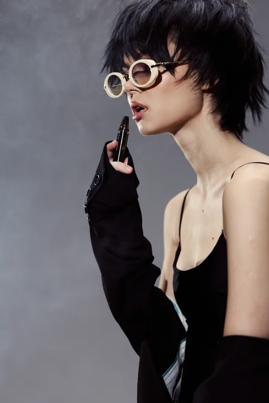 Stylish woman wearing round sunglasses and black outfit with edgy hairstyle posing thoughtfully against gray background