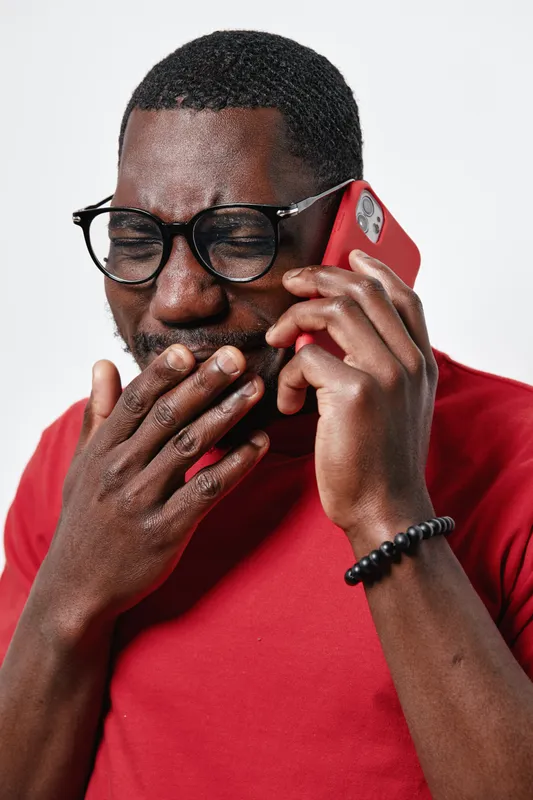 African American man with dark skin wearing glasses and red shirt talking on smartphone covering mouth with hand expressing surprise or shock. Closeup portrait on white background.