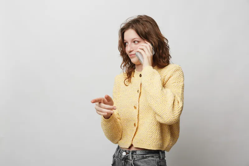 Young woman in yellow knitted sweater talks on smartphone and points aside. Casual style, communication, and technology concept with neutral background and natural lighting.