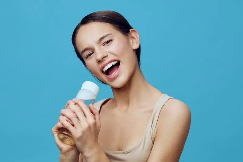 Young woman with a joyful expression holding a bottle of beverage against a light blue background, representing hydration and healthy lifestyle