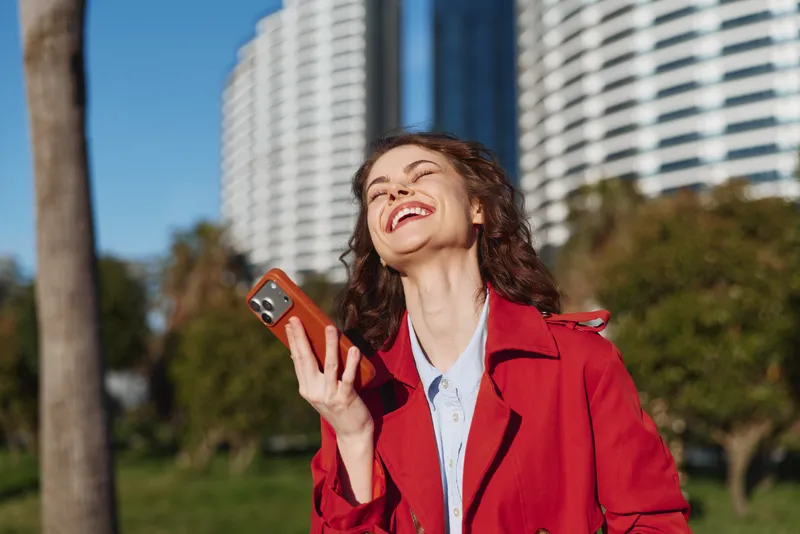 A smiling woman in a redcoat holds a phone and gazes upward in a sunny urban park with a tall skyscraper backdrop, capturing a moment of joyful connection and energy.