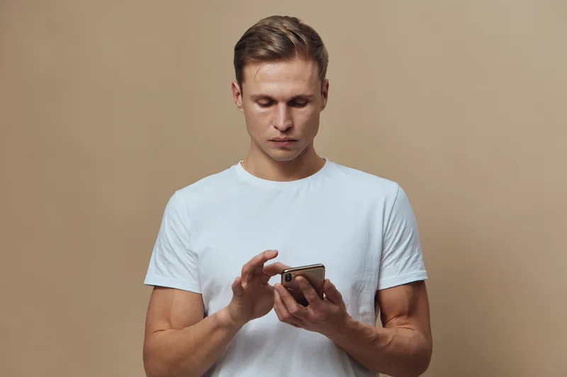 Modern young man in plain white t-shirt using smartphone device holding in hand isolated on beige background studio portrait digital technology concept