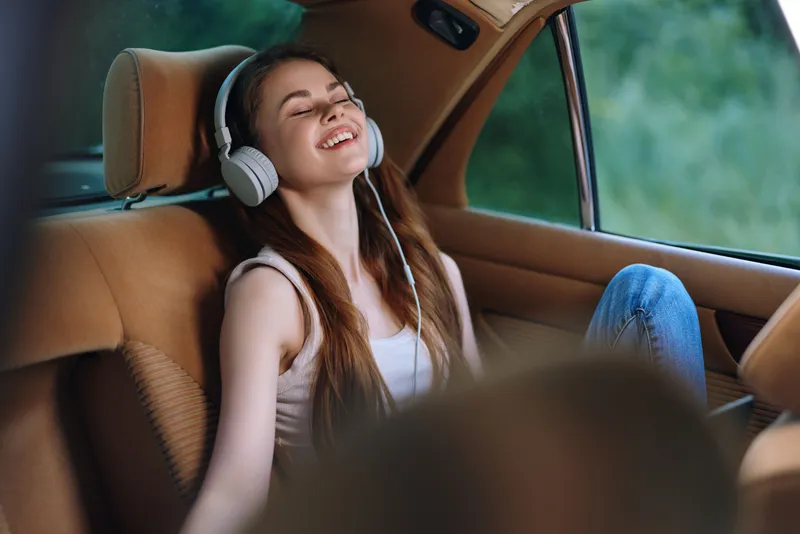 Young woman enjoying music in a car, wearing headphones, relaxing with a smile, natural background, representing joy and serenity.