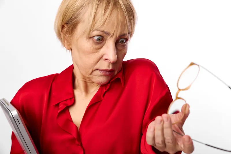 Elderly woman in red blouse, surprise on her face, holding glasses beside a laptop, indoor white background, soft daylight, worried about vision and screen focus today