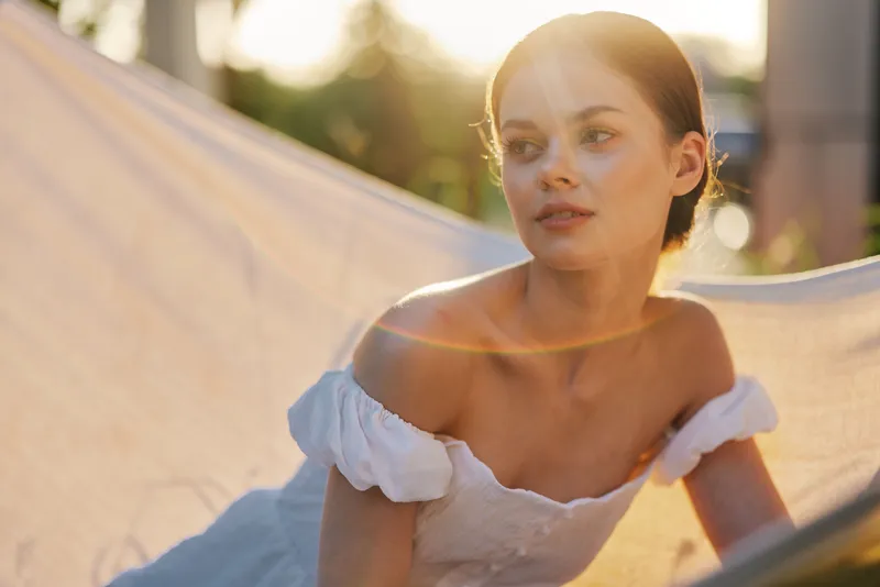 A serene young woman in a white dress enjoys a sunlit moment in a hammock, radiating tranquility and elegance in a lush garden setting