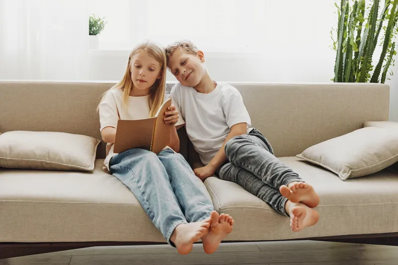 Two barefoot children sitting on beige sofa reading a book together in bright living room. Concept of sibling bonding, childhood learning, family time and casual home environment.