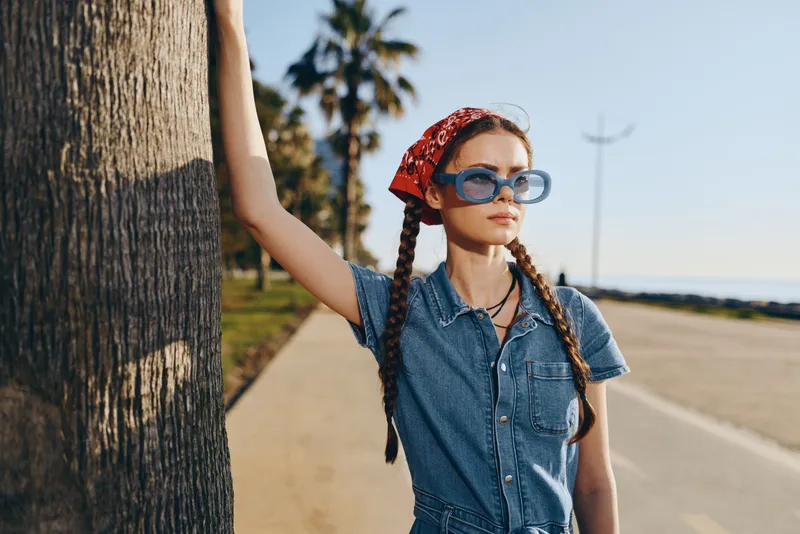 Lifestyle woman in modern boho-western denim street style wearing blue sunglasses and red bandana, standing by palm tree on sunny urban street with warm film color tones.
