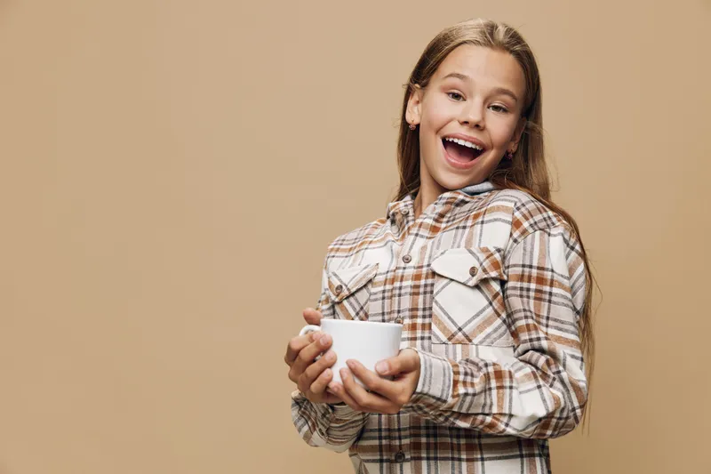 Girl holding white cup smiling and looking away on beige background. Young female with long hair in plaid shirt enjoying beverage in studio portrait.