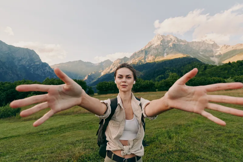 Cheerful woman outdoors reaches toward the camera with open arms, mountain meadow scene, sunlit day, casual outfit, sense of adventure, natural colors, and positive mood