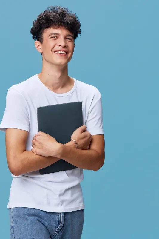 smiling man holding laptop, casual young adult in white t-shirt and jeans, standing against solid blue background with relaxed confident posture