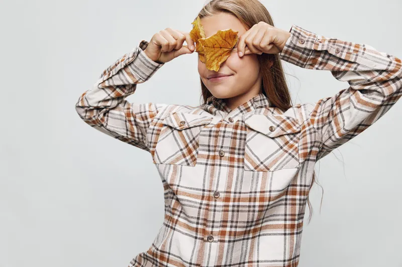 Girl in plaid shirt covering eyes with autumn leaf, smiling and posing isolated on white background for seasonal concept and childhood fun outdoors.